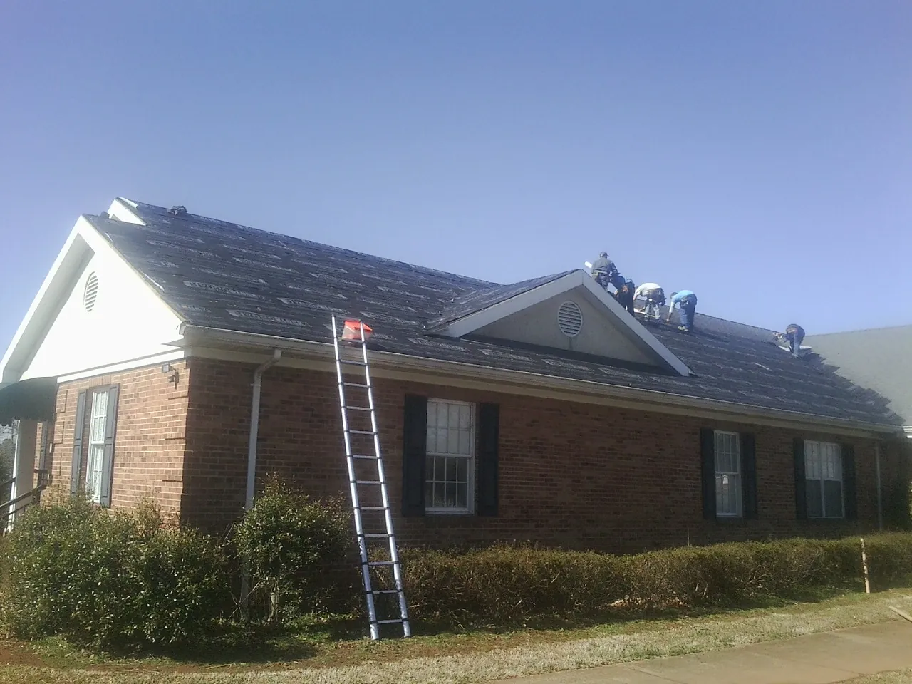 Crew tearing shingles off a brick ranch roof with a ladder set — Metal Roof Repair work by our crew in Champlain