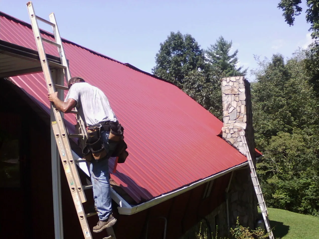 Roofer on a ladder beside a red metal roof and stone chimney — Soffit & Fascia Repair by Roofing Brothers in Champlain